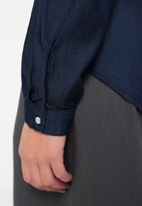 Camisa de ganga em azul escuro, com mangas ajustadas e punhos com botão. Usada com uma peça de roupa cinza de textura suave.