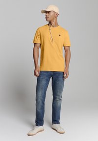 Geel katoenen t-shirt met korte mouwen, gecombineerd met blauwe denim jeans. Model draagt een beige pet en lichte sneakers met een gestructureerde zool.