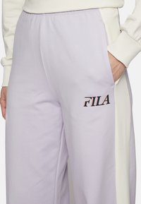 Lichtpaarse joggingsbroek met een witte zijpaneel, voorzien van een zwart "FILA" logo aan de voorkant. Gemaakt van zachte stof met een elastische tailleband.