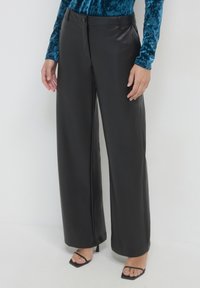 Donna che indossa pantaloni neri a gamba larga in similpelle, una maglietta a maniche lunghe in velluto color turchese e sandali neri con tacco a punta quadrata, in piedi su uno sfondo bianco.