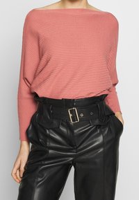 Maglione rosa a coste con scollo a barca abbinato a pantaloni neri in pelle a vita alta con cintura larga e tasche visibili.