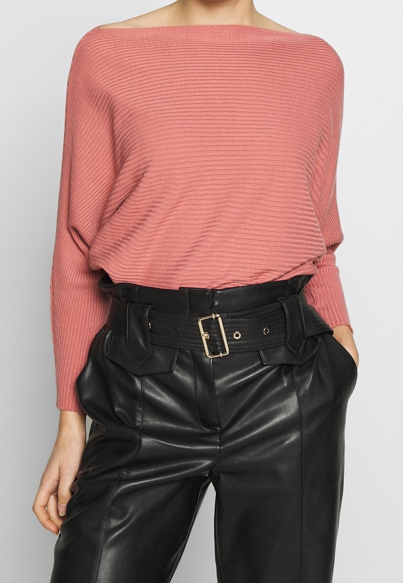 Maglione rosa a coste con scollo a barca abbinato a pantaloni neri in pelle a vita alta con cintura larga e tasche visibili.