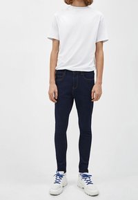 Vit t-shirt med kort ärm, mörkblå skinny jeans och vita sneakers med blå snören. Enkelt, avslappnat outfit mot en neutral bakgrund.