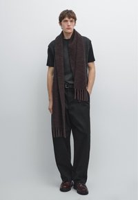 Jongeman in zwarte broek, zwart t-shirt, grijze mouwloze sweater, lange donkerbruin sjaal en donkerbruine schoenen, staand tegen een lichte muur.