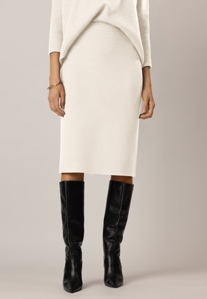 Woman wearing a cream-colored knit skirt, matching sweater, and black knee-high leather boots against a neutral background.