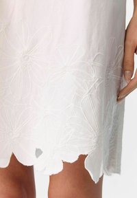 Abito bianco con ricami floreali all'orlo, bordo scallopato e una texture liscia. Primo piano di intricati modelli floreali e contorni.