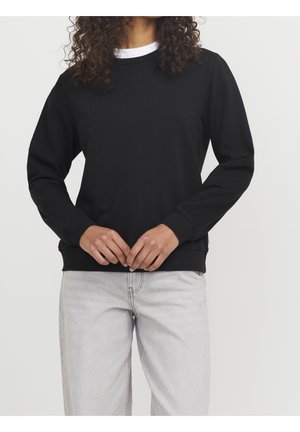 Osoba z kręconymi włosami, ubrana w czarną bluzę z długim rękawem na białej koszulce oraz jasnoszare dżinsy, stojąca z rękami splecionymi.