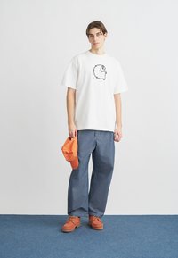 Λευκό βαμβακερό T-shirt με μαύρο γραφικό σχέδιο, συνδυασμένο με μπλε τζιν παντελόνι, πορτοκαλί καπέλο και ταμπά παπούτσια με κορδόνια σε μπλε δάπεδο.