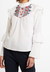 Vit blus med volangärmar och broderat blommönster i rött och blått, med en strukturerad prickdesign på tyget.