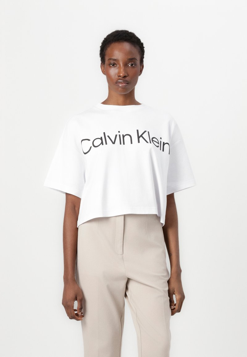 Kuttet hvit t-skjorte med "Calvin Klein" trykket i svart. Korte ermer, myk bomullsduk, avslappet passform. Sammen med beige bukser.