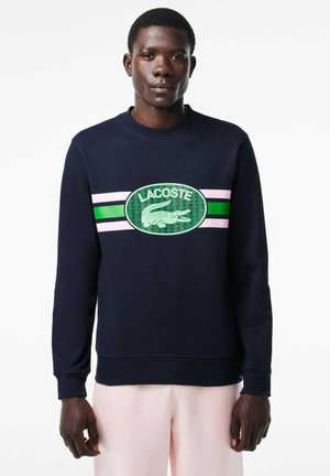 Granatowy sweatshirt z okrągłym dekoltem, z zielono-różowym paskowanym wzorem graficznym i dużym zielonym logo krokodyla na środku.