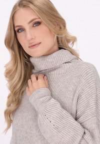 Hellgrauer Rollkragenpullover mit gerippter Textur und langen Ärmeln. Verfügt über einen umgeschlagenen Kragen und ein kleines Lochdetail am Ärmel.