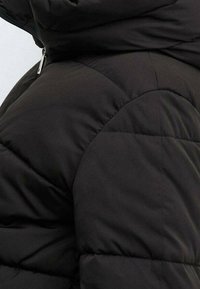 Gros plan sur l'épaule et le haut du bras d'une veste matelassée noire avec une fermeture éclair visible sur le col.