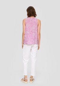 Blusa senza maniche floreale viola con colletto alto e dettagli con apertura a chiave sul retro, abbinata a pantaloni di lino bianchi e sandali beige.
