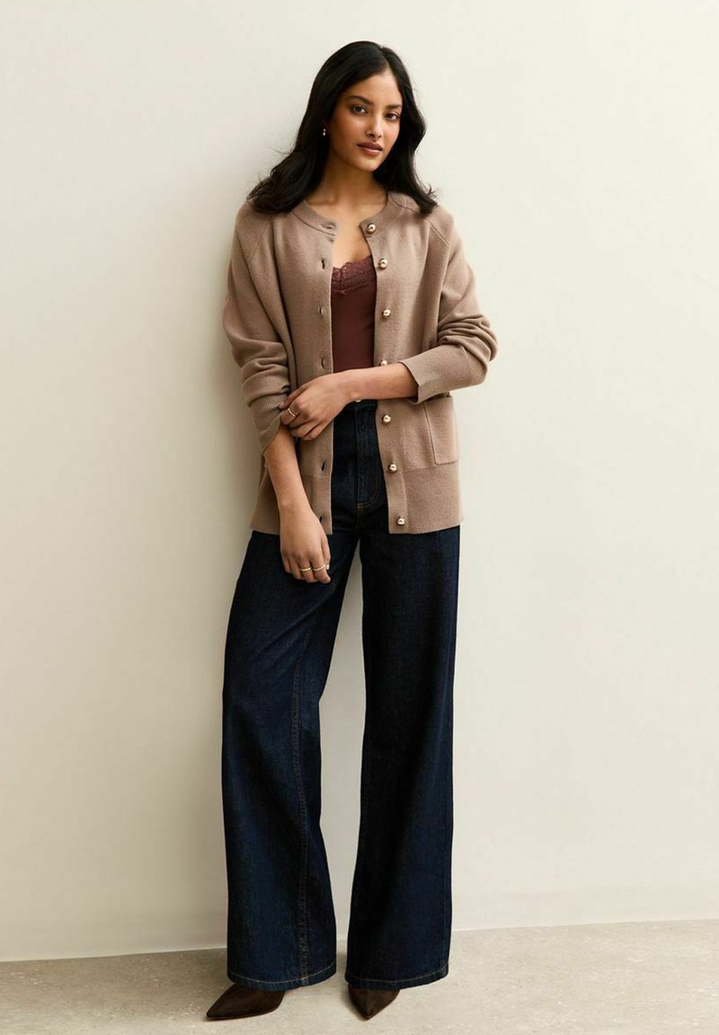 Cardigan de punto beige con botones de perla, llevado sobre una camiseta sin mangas oscura. Combinado con jeans de pierna ancha y zapatos marrones de punta.