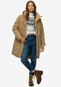 Vrouw in een beige gewatteerde winterjas met capuchon, een gebreid patroontrui, een blauwe spijkerbroek, een bruine riem en bruine veterschoenen, staand met één been gekruist.