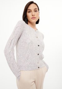Cardigan grigio chiaro a maglia con collo tondo, dotato di cinque bottoni metallici e polsini a coste, indossato sopra pantaloni beige.