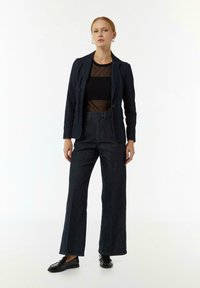 Dunkelblauer Jeansblazer mit taillierter Passform, über einem transparenten schwarzen Oberteil getragen und kombiniert mit weit geschnittenen dunklen Jeans. Schwarze Loafers runden das Outfit ab.