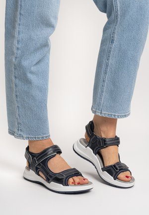Feet wearing open-toe black and white sandals with adjustable straps, paired with light blue denim jeans against a plain background.