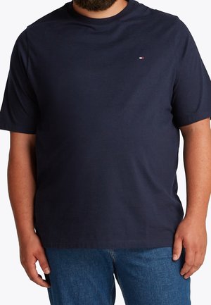 Homme portant un t-shirt bleu marine uni à manches courtes avec un petit logo rouge, blanc et bleu sur la poitrine gauche et un jean bleu.
