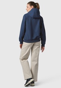 Mörkblå huvjacka med elastiska ärmslut och nederkant, i kombination med ljusbeiga breda byxor och svarta sneakers, sedd bakifrån.