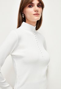 Witte langslieve top met een hoge kraag, voorzien van een knoopdetail aan de voorkant. Materiaal lijkt glad met een licht gestructureerde afwerking.
