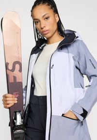 Smučarska jakna v svetlo sivi in temno sivi barvi, z zadrgo spredaj, nastavljivo kapuco in več žepi. Roka drži smučko z grafičnimi vzorci.