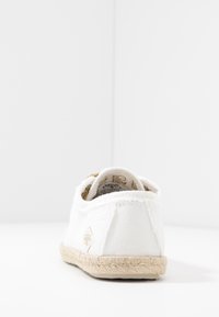 Bílé plátěné tenisky s jutu obalenou podrážkou, se strukturovaným zadním dílem, zlatými akcenty uvnitř a švy podél okrajů.