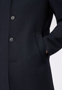 Cappotto di lana navy con tessuto testurizzato, quattro bottoni neri e dettagli delle tasche laterali per un aspetto sartoriale.