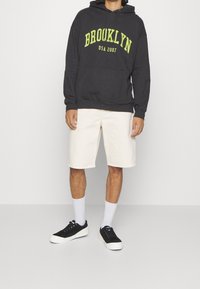 Sudadera con capucha gris oscuro con el texto "BROOKLYN USA 2007" en amarillo brillante, combinada con pantalones cortos beige claro. Zapatillas negras y calcetines blancos completan el conjunto.