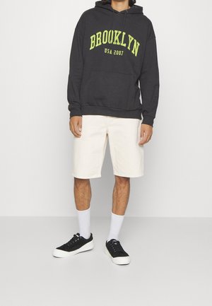 Tmavě šedá mikina s kapucí s nápisem „BROOKLYN USA 2007“ ve světlé žluté barvě, doplněná světle béžovými šortkami. Černé tenisky a bílé ponožky dokončují outfit.