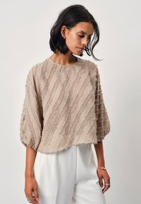 Beige, leichtes Oberteil mit lockerer Passform und strukturiertem, welligem Muster. Hat einen runden Ausschnitt und kurze, weite Ärmel. Kombiniert mit weißen Hosen.