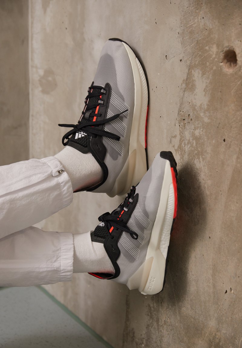 Graue Sportschuhe mit schwarzen Akzenten, Mesh-Einsätzen, roten Details und dicken weißen Sohlen, vor einem Betonhintergrund platziert.