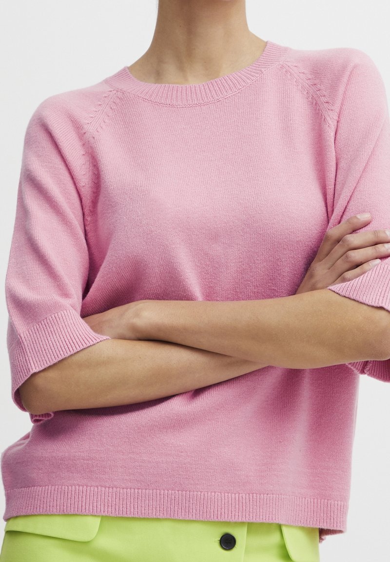 Personne croisant les bras, portant un pull en tricot à manches courtes rose pastel et un bas vert citron avec un bouton noir.