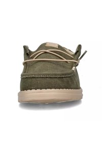 Zapatilla de lona verde oliva con cordones beige, punta redonda, acentos cosidos y suela de goma texturizada. Presenta una lengüeta etiquetada.