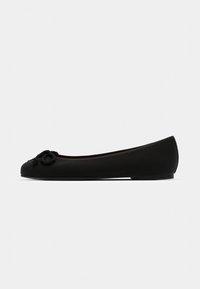Chaussure ballerine noire avec bout rond et détail de nœud décoratif à l’avant, vue de profil sur fond uni.