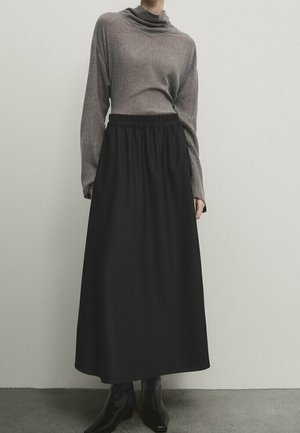 Personne portant un haut gris à manches longues, une jupe noire taille haute en A et des bottines noires sur un fond clair uni.