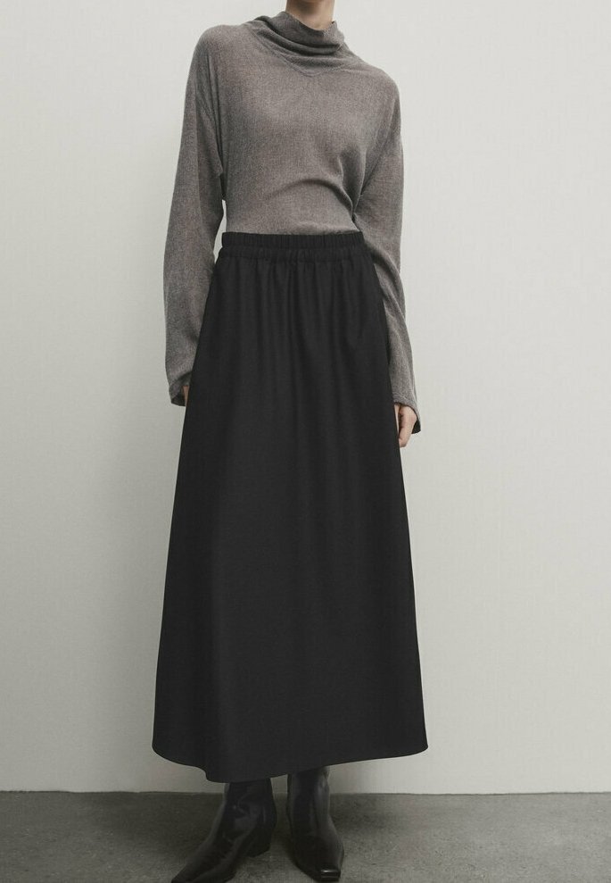 Personne portant un haut gris à manches longues, une jupe noire taille haute en A et des bottines noires sur un fond clair uni.