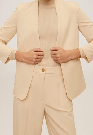Mains ajustant un blazer beige par-dessus un pull à col montant côtelé beige, assorti à un pantalon beige sur mesure avec une fermeture à bouton.