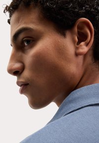 Profilový pohled na osobu s kudrnatými vlasy, která má na sobě texturovanou modrou košili. Významné rysy zahrnují výraznou čelist a hladkou pleť.