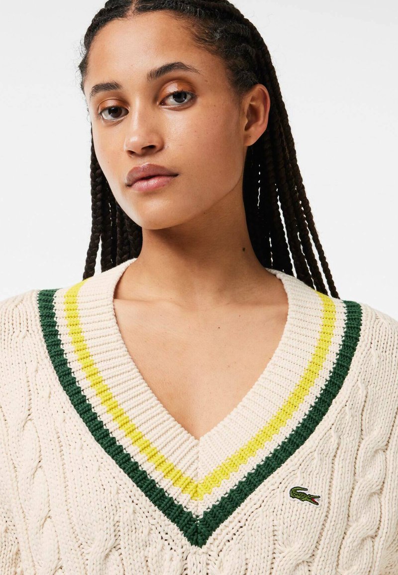 Pull en tricot torsadé crème avec un col en V, présentant des rayures vertes et jaunes le long du col. Petit logo brodé sur la poitrine.