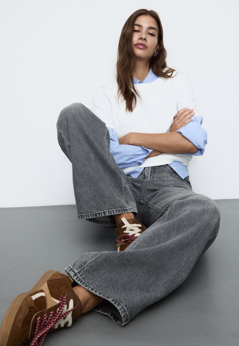 Jeans in denim grigio a gamba larga con orlo sfrangiato, abbinati a una felpa bianca indossata sopra una camicia a strisce azzurre. Sneakers marroni con lacci unici.