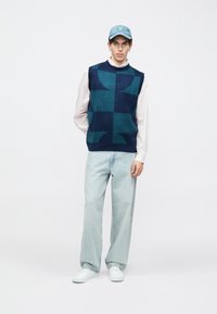 Μπλε ναυτικό πλεκτό αμάνικο πουλόβερ με γεωμετρικό μοτίβο, συνδυασμένο με ανοιχτόχρωμα τζιν και λευκά παπούτσια, ολοκληρωμένο με ανοιχτό μπλε καπέλο.