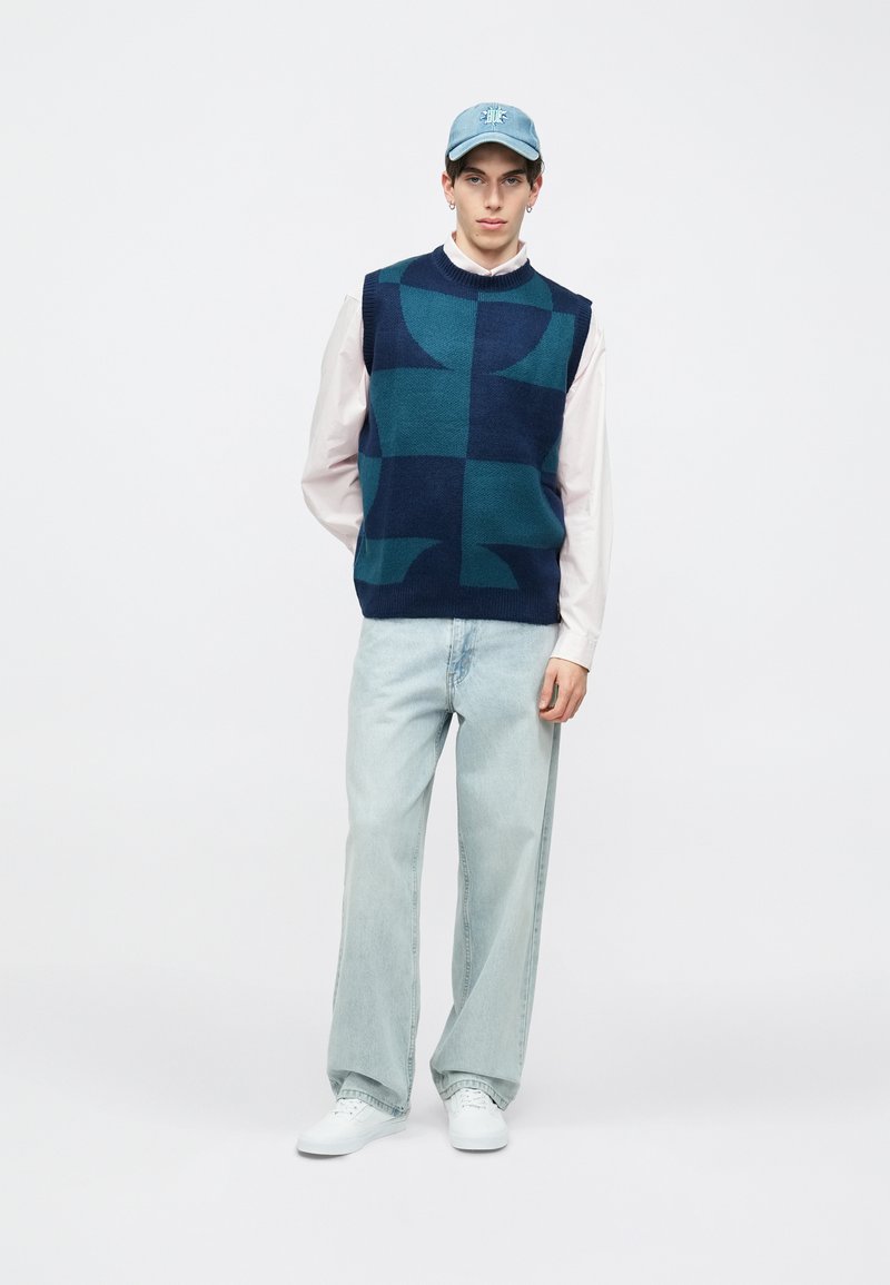 Μπλε ναυτικό πλεκτό αμάνικο πουλόβερ με γεωμετρικό μοτίβο, συνδυασμένο με ανοιχτόχρωμα τζιν και λευκά παπούτσια, ολοκληρωμένο με ανοιχτό μπλε καπέλο.