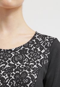 Svart topp med en vit spetspanel på den övre framsidan, rund halsringning och en texturerad tyg med långa ärmar.