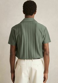 Cămașă cu mânecă scurtă, de culoare verde măslinie, cu dungi verticale, cu guler și croială lejeră. Pur tată cu pantaloni de culoare crem, cu două buzunare pe spate.
