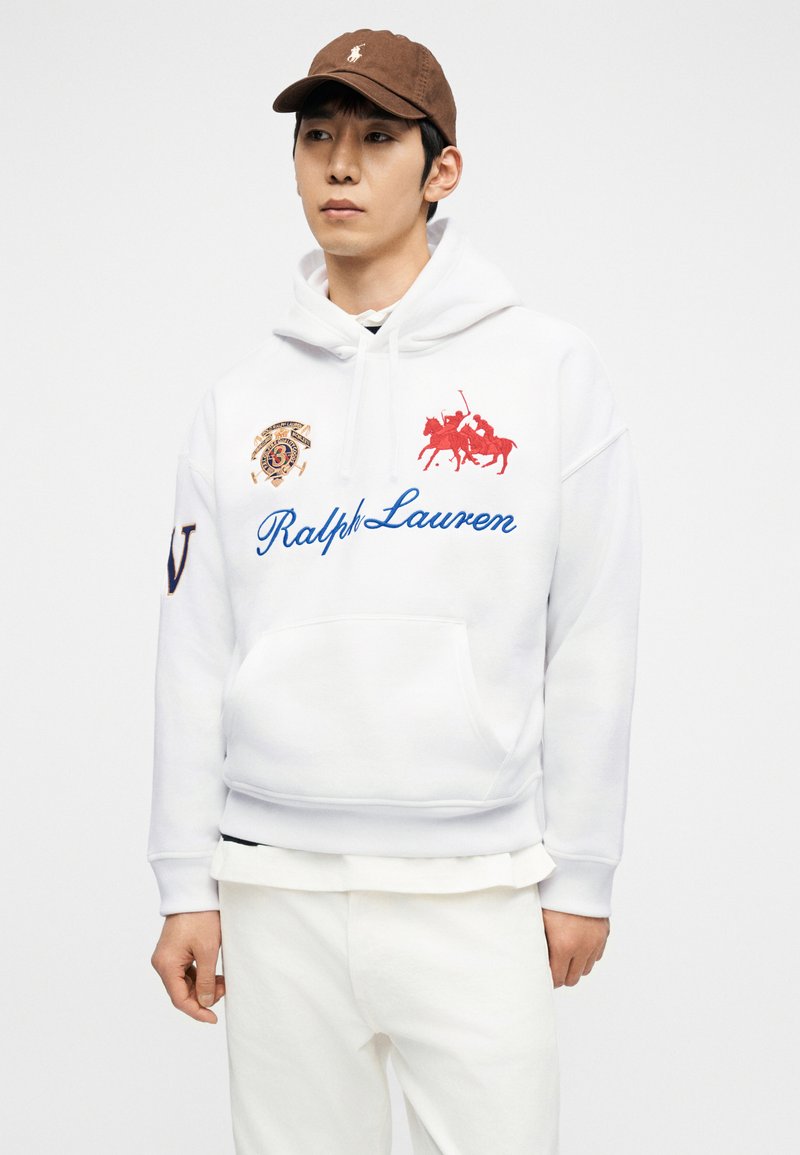 Homme portant un sweat à capuche blanc Ralph Lauren avec des logos bleus et rouges, un pantalon blanc et une casquette de baseball marron, debout devant un fond blanc.