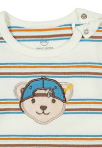 Chemise à manches courtes rayée en crème, bleu, brun et orange. Présente un ours brodé portant une casquette bleue avec une accentuation jaune.