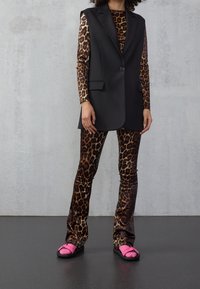 Černá šitá vesta přes top s leopardím potiskem a odpovídajícími kalhotami; má lesklou tkaninu, přiléhavý design a růžové sandály s černou podrážkou.