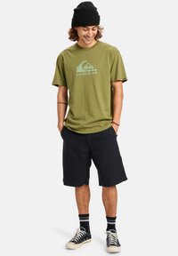 Olivově zelené bavlněné tričko s logem Quiksilver, spárované s černými šortkami, černými teniskami a pruhovanými ponožkami. Krátké rukávy a kulatý výstřih.